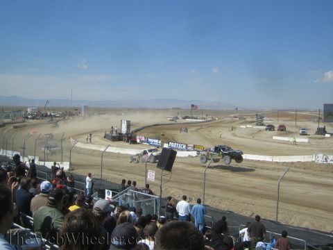 1990 Ford Bronco - Spectators watch off-road race trucks kicking up dust on a dirt short-course oval in a dry desert setting.