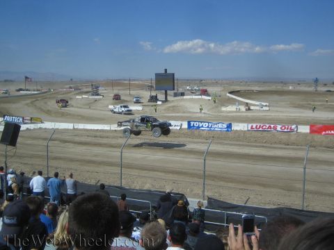 1990 Ford Bronco - off-road race truck launches off a dirt jump as a crowd watches from the grandstand, Toyo Tires and Lucas Oil banners behind.