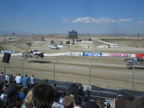 1990 Ford Bronco - white off-road race truck lands a jump on a dirt course ringed with sponsor banners and a watching crowd.