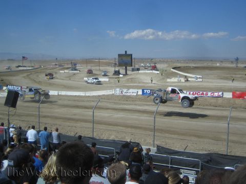 1990 Ford Bronco - off-road race truck catches air beside a Lucas Oil banner on a dusty dirt short-course track.