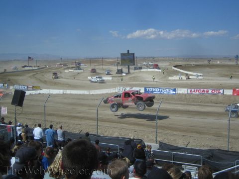 1990 Ford Bronco - red off-road race truck launches off a jump on a dirt short-course track while spectators look on.