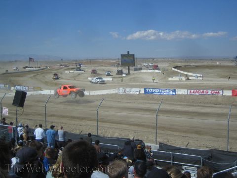 1990 Ford Bronco - orange off-road race truck catches air on a dusty dirt course ringed with sponsor banners and a crowd.