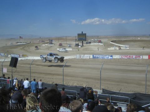 1990 Ford Bronco - white off-road race truck launches a jump on a dusty dirt track as a grandstand crowd watches.