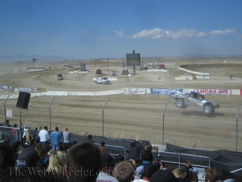1990 Ford Bronco - silver off-road race truck catches air on a dirt short-course track lined with sponsor banners.