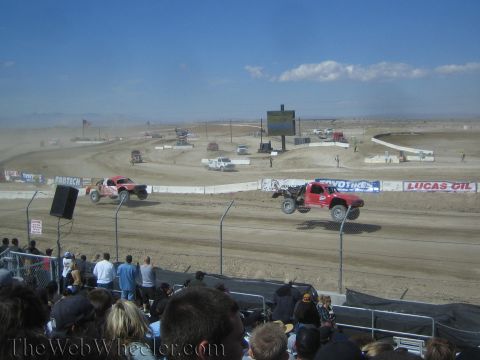 1990 Ford Bronco - Two off-road race trucks battle on a dusty dirt short-course track as spectators watch from the stands.
