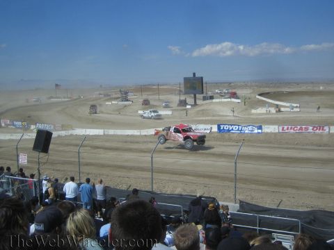 1990 Ford Bronco - off-road race truck races along a dirt short-course track with a hazy desert backdrop and watching crowd.