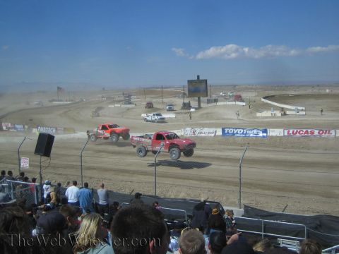 1990 Ford Bronco - Two off-road race trucks race together on a dusty dirt short-course track as a crowd looks on.
