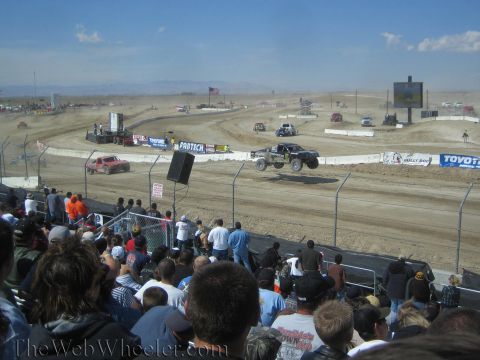 1990 Ford Bronco - Off-road race trucks navigate a dirt short-course track as a large grandstand crowd watches.