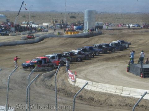 1990 Ford Bronco - row of off-road race trucks is staged in a line on a dirt short-course track before a desert race.
