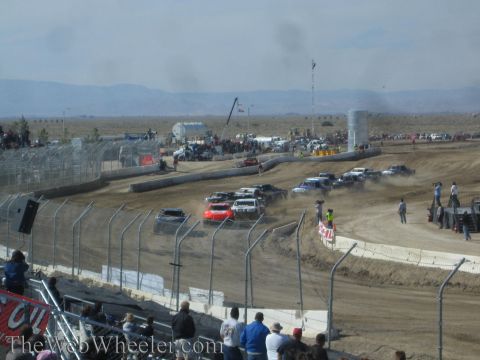1990 Ford Bronco - pack of off-road race trucks bunches through a turn on a dusty dirt short-course track with mountains behind.