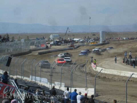 1990 Ford Bronco - Off-road race trucks race through a dusty turn on a dirt short-course track with a desert mountain backdrop.