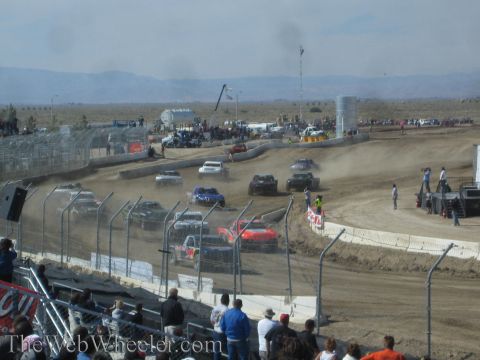 1990 Ford Bronco - field of off-road race trucks races across a dusty dirt short-course track with mountains in the distance.