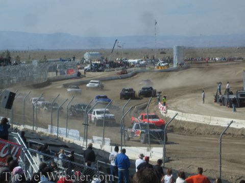 1990 Ford Bronco - Off-road race trucks descend through a dusty turn on a dirt short-course track as spectators watch.