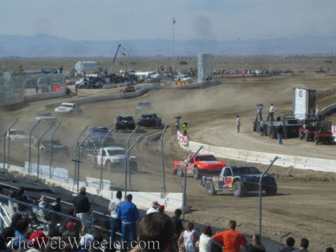 1990 Ford Bronco - Off-road race trucks round a dusty turn on a dirt short-course track with a desert backdrop and crowd.