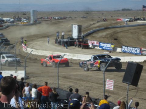 1990 Ford Bronco - Red Bull-liveried off-road race truck leads others through a turn on a dirt short-course track past sponsor banners.