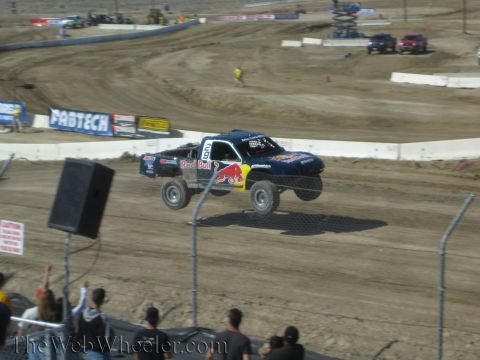 1990 Ford Bronco - dark blue Red Bull-liveried off-road race truck launches off a jump on a dirt short-course track.