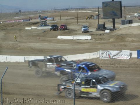 1990 Ford Bronco - Stadium-style off-road trucks race nose-to-tail along a dirt track lined with concrete barriers, a Bully Dog banner and large video screen…