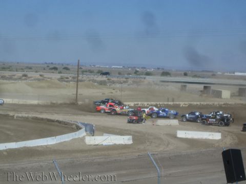 1990 Ford Bronco - cluster of brightly liveried short-course trucks bunch together on a dirt track with a flat desert and overpass in the background.
