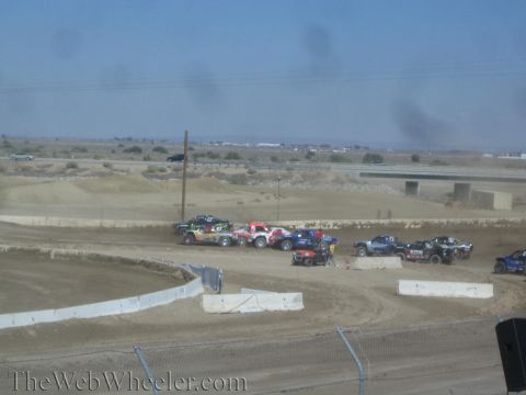 1990 Ford Bronco - Several short-course race trucks jostle in a tight pack on a dusty dirt track under a hazy desert sky.