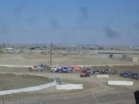 1990 Ford Bronco - field of short-course trucks works through a turn on a dirt track, with desert scrub and a highway overpass beyond.