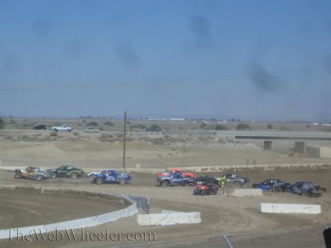 1990 Ford Bronco - spread-out line of liveried short-course trucks races across a wide dirt track flanked by white barriers in the desert.