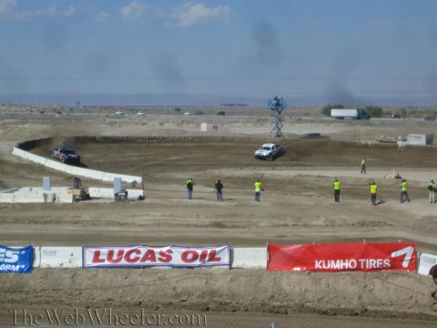 1990 Ford Bronco - single off-road truck rounds a banked dirt turn while spectators and crew stand near Lucas Oil and Kumho Tires trackside banners.