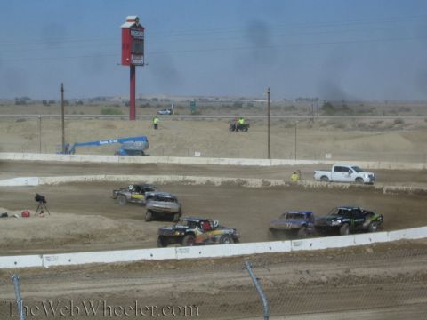 1990 Ford Bronco - Four short-course race trucks run along a dirt straight, a tall red sign and white pickup chase truck in the background.