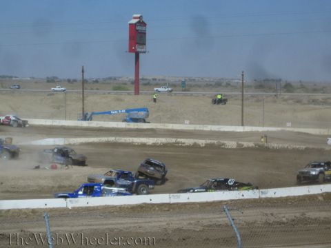 1990 Ford Bronco - Short-course trucks fan out and throw dust on a dirt track, with a blue lift and tall red sign behind the barrier wall.