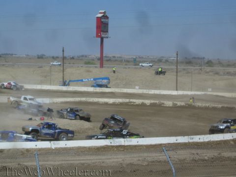 1990 Ford Bronco - One short-course truck lifts off the dirt while rivals battle around it on a desert track marked by a tall red sign.