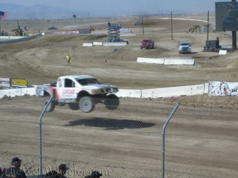 1990 Ford Bronco - white Lucas Oil short-course truck launches over a jump on a dirt track as spectators watch from behind the fence.