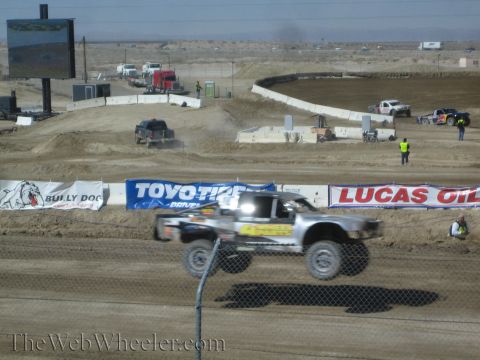 1990 Ford Bronco - silver short-course truck catches air over a dirt jump in front of Bully Dog, Toyo Tires and Lucas Oil trackside banners.