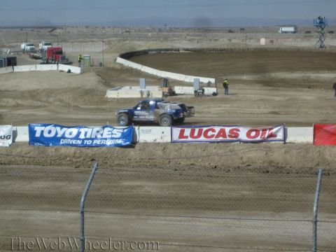 1990 Ford Bronco - blue short-course truck races along a dirt track behind Toyo Tires and Lucas Oil banners in a wide desert venue.