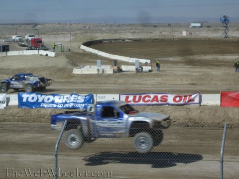 1990 Ford Bronco - silver-and-blue short-course truck leads a blue rival down a dirt straight lined with Toyo Tires and Lucas Oil banners.