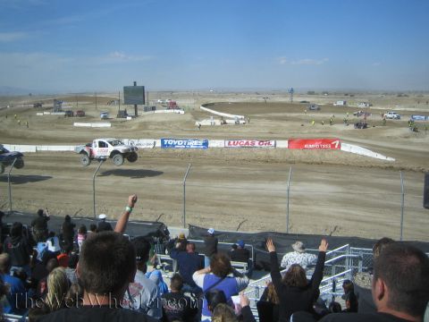 1990 Ford Bronco - grandstand crowd, some with arms raised, watches short-course trucks race past Lucas Oil and Kumho Tires banners at a desert track.