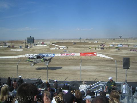 1990 Ford Bronco - short-course truck catches air over a dirt jump as a packed grandstand watches in a wide desert racing complex.