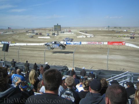 1990 Ford Bronco - Spectators in a grandstand watch a short-course truck launch off a jump past Toyo Tires and Lucas Oil banners at a desert track.