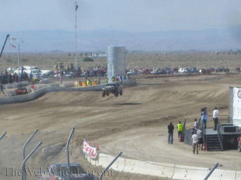 1990 Ford Bronco - short-course truck catches air near a large metal tank as crowds line the fenced dirt track in a dusty desert venue.