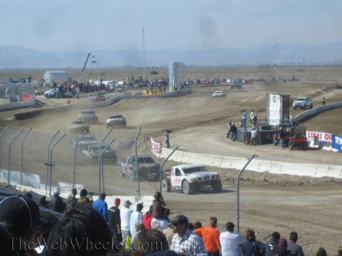 1990 Ford Bronco - field of short-course trucks streams along a winding, dusty dirt course as spectators watch from behind the fence.