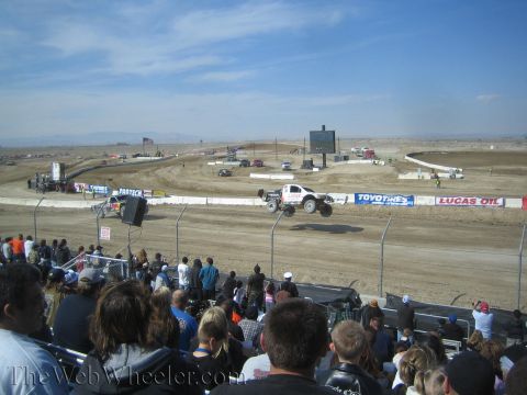 1990 Ford Bronco - white short-course truck catches air over a dirt jump in front of a grandstand crowd, with Toyo Tires and Lucas Oil banners behind.