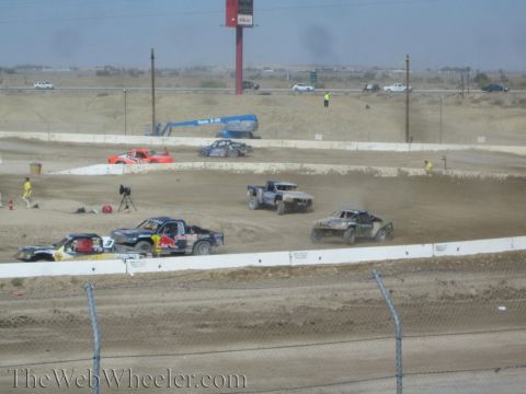 1990 Ford Bronco - Several short-course trucks, including a Red Bull-liveried truck, battle through a dirt turn under a tall red sign.