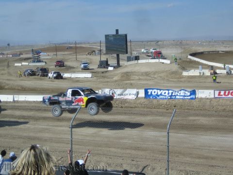 1990 Ford Bronco - Red Bull-liveried short-course truck catches air over a dirt jump beside Bully Dog and Toyo Tires banners as fans cheer.