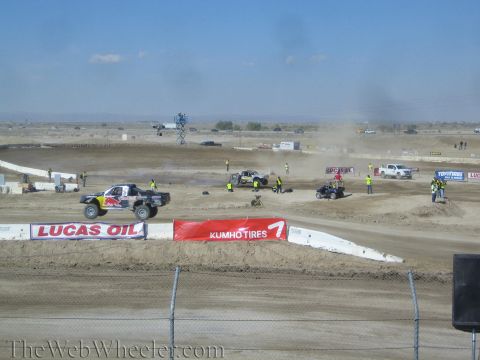 1990 Ford Bronco - white off-road race truck rounds a desert dirt course lined with Lucas Oil and Kumho Tires sponsor banners as crews work the infield.