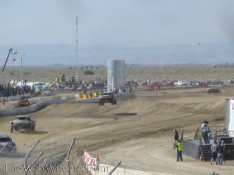 1990 Ford Bronco - Off-road race trucks kick up dust on a dirt track in a desert valley, with a tall light tower, water tank, and roadside crowd in the backgr…