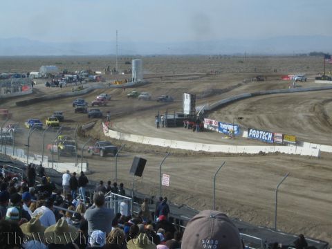 1990 Ford Bronco - grandstand crowd watches off-road race trucks circle a dusty dirt oval lined with Fabtech and other sponsor banners under a hazy desert sky.