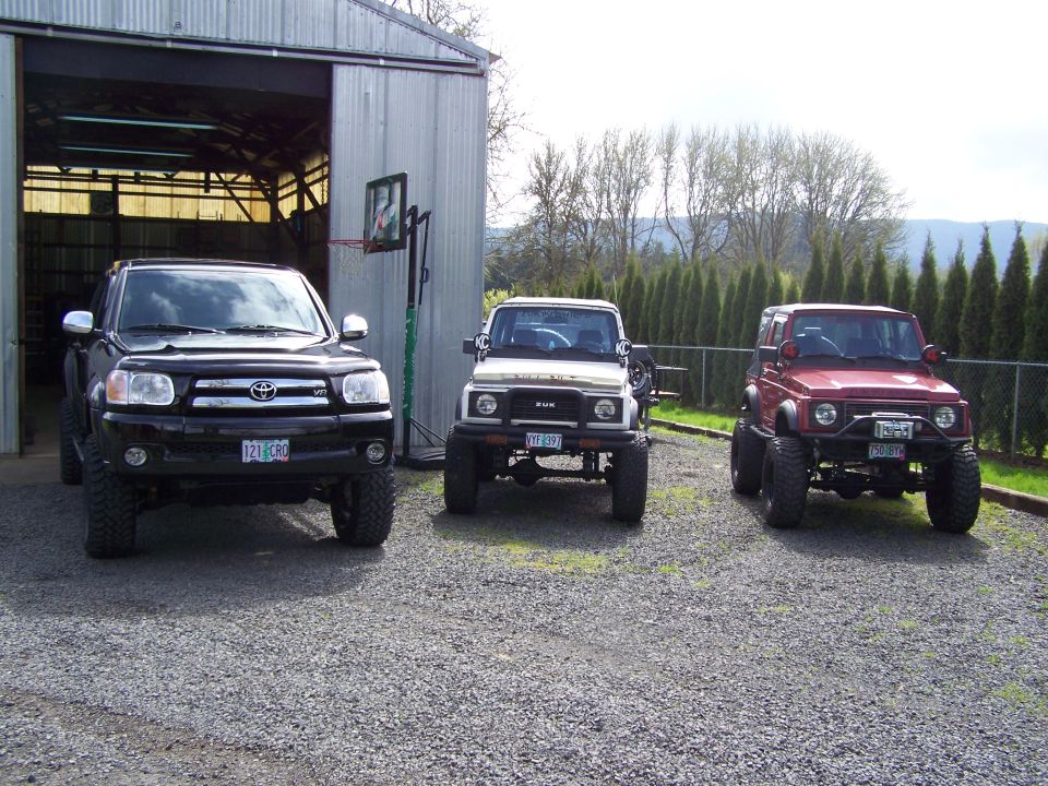 Black Toyota and two lifted Suzuki Samurais on gravel by a metal barn