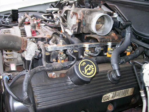 Engine bay with oil cap, hoses, and wiring. Part number visible on black cover