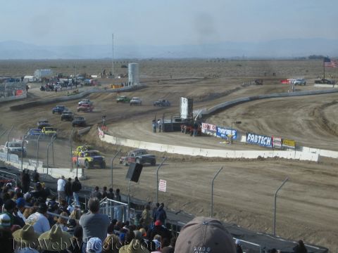 1990 Ford Bronco - Off-road race trucks run a banked dirt oval framed by Fabtech banners and a US flag, seen from the stands over a crowd of spectators.