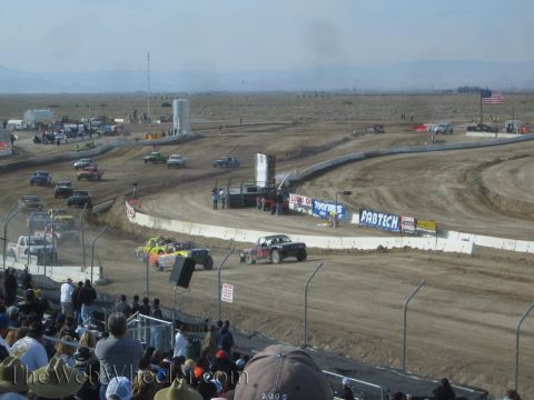 1990 Ford Bronco - Several off-road race trucks navigate a dusty dirt oval lined with sponsor banners, viewed from a packed grandstand in the desert.