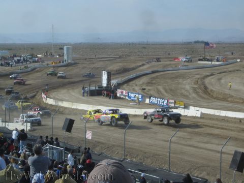 1990 Ford Bronco - pack of off-road race trucks corners on a dusty dirt oval lined with Fabtech and other banners, with grandstand spectators in the foregroun…