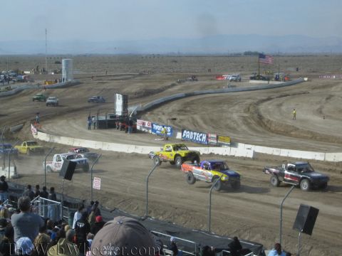 1990 Ford Bronco - Off-road race trucks battle through thick dust on a dirt oval lined with sponsor banners and a US flag as spectators watch from the stands.